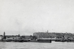 Die Aufnahme zeigt eine befestigte Hafenanlage mit Molen, Kränen und militärischen Einrichtungen vor einer Festung auf einem Hügel. Im Vordergrund sind Hafenbecken und Pier-Anlagen der Kaiserlichen Marine zu erkennen.