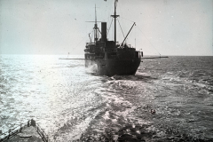Ein deutsches Kriegsschiff mit charakteristischem Schornstein und Masten fährt auf ruhiger See. Die Aufnahme wurde vom Deck des U-Bootes S.M. U 39 aus gemacht, erkennbar am Reling im Vordergrund.