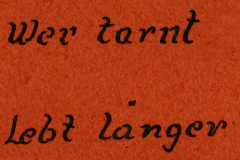 Handschriftliche Inschrift auf rotem Untergrund: 'Wer tarnt lebt länger'