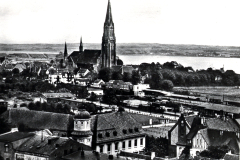 Stadtansicht mit neugotischer Kirche und Eisenbahnanlage an Flusslandschaft