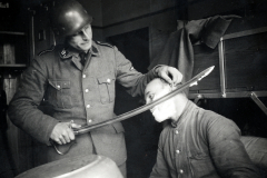 SS-Offizier zeigt Waffe an sitzendem Mann in Uniform